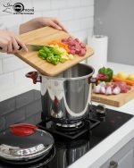 O.M.S/ONEMORE 14-liter stainless steel pressure cooker with red handles sitting on a kitchen stovetop, featuring an open lid with steaming hot meat stew inside.