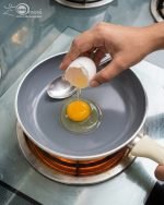 Falez 9-piece cream colored non-stick cookware set displayed on a wooden table, featuring a granite ceramic interior, glass lids with blue knobs, and a frying pan, made in Turkey.