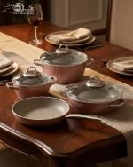 Falez 9-piece pink ceramic granite cookware set featuring a fry pan tossing vegetables, a shallow casserole, and three pots with glass lids, arranged on a kitchen counter and dining table.