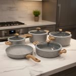 Falez 9-piece grey granite non-stick cookware set displaying a frying pan, shallow casserole, and deep pots with glass lids and wooden-style handles on a kitchen countertop.