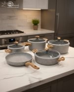 Falez 9-piece grey granite non-stick cookware set displaying a frying pan, shallow casserole, and deep pots with glass lids and wooden-style handles on a kitchen countertop.