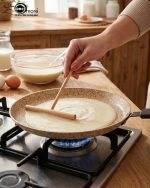 Falez non-stick granite crepe pan in beige speckled finish, shown cooking a golden-brown crepe on a gas stove with a wooden spreader. Option 2: Person flipping a crepe in a Falez granite coated pan, demonstrating non-stick surface and low-oil cooking capabilities.
