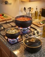 One More 12-piece black granite non-stick cookware set displayed on a wooden table, featuring gold handles, glass lids, a fry pan, shallow casserole, deep pots, and cooking spoons.