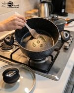 One More 24cm black granite non-stick cooking pot with glass lid and heat-resistant silicone handles, shown preparing healthy low-oil food.