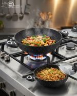 One More black granite non-stick wok set stacked in four sizes (24cm, 26cm, 28cm, 30cm) with glass lids and double handles, featuring a speckled texture for healthy low-oil cooking."