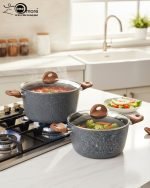 Three-piece One More non-stick granite cooking pot set in grey speckled texture, featuring 24cm, 26cm, and 28cm pots with glass lids and wood-effect silicone handles, displayed on a modern kitchen counter.