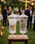 A luxurious 4.5-liter crystal-cut glass beverage dispenser by One More, featuring an intricate diamond pattern and silver spigot, displayed on a formal dining table with lemon-infused water.