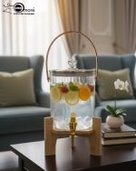 A One More brand 3.6 liter ribbed glass water dispenser filled with lemon slices and ice, featuring a gold tap, a brown leather handle, and a heart-shaped lid knob, resting on a light wood bamboo stand in a modern kitchen and living room setting.