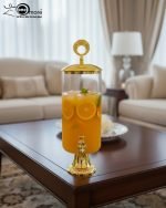 Elegant 3.5L clear glass water dispenser with luxury gold base, gold lid with ring handle, and a gold spigot, filled with infused water and citrus slices on a marble kitchen countertop.
