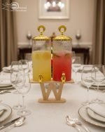 A luxury dual glass beverage dispenser set by One More featuring a 3.3L smooth jar and a 3.3L hammered glass jar, equipped with gold metallic spigots and lids, displayed on a stylish W-shaped wooden stand on a decorated dinner table.