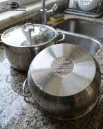 Stacked collection of Falez stainless steel cooking pots in seven sizes ranging from 18cm to 30cm, featuring glass lids and a polished silver finish, displayed on a wooden table for the One More kitchenware brand.