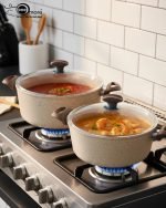 Falez 9-piece beige granite non-stick cookware set arranged on a kitchen counter, featuring a fry pan, shallow casserole, and three deep pots with glass lids, showcasing healthy low-oil cooking.