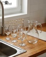 Pasabahce 7-piece glass water set featuring one clear jug and six stemmed glasses with elegant gold rims, displayed on a wooden surface.