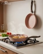 Falez 9-piece pink granite non-stick cookware set including 3 cooking pots with lids, 1 shallow casserole with lid, and 1 frying pan displayed on a kitchen counter and dining table, demonstrating healthy low-oil cooking with steam rising from hot food.