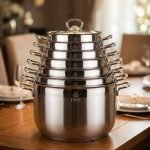 Stacked collection of Falez stainless steel cooking pots in seven sizes ranging from 18cm to 30cm, featuring glass lids and a polished silver finish, displayed on a wooden table for the One More kitchenware brand.