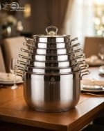 Stacked collection of Falez stainless steel cooking pots in seven sizes ranging from 18cm to 30cm, featuring glass lids and a polished silver finish, displayed on a wooden table for the One More kitchenware brand.