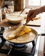 Falez non-stick granite crepe pan in beige speckled finish, shown cooking a golden-brown crepe on a gas stove with a wooden spreader. Option 2: Person flipping a crepe in a Falez granite coated pan, demonstrating non-stick surface and low-oil cooking capabilities.