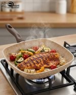 Falez square granite non-stick grill pan cooking grilled chicken breast and vegetables with perfect char marks, featuring a beige speckled finish and ergonomic brown handle.