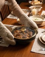 One More 12-piece black granite non-stick cookware set displayed on a wooden table, featuring gold handles, glass lids, a fry pan, shallow casserole, deep pots, and cooking spoons.