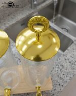 A luxury dual glass beverage dispenser set by One More featuring a 3.3L smooth jar and a 3.3L hammered glass jar, equipped with gold metallic spigots and lids, displayed on a stylish W-shaped wooden stand on a decorated dinner table.