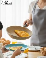 Falez 9-piece beige granite non-stick cookware set arranged on a kitchen counter, featuring a fry pan, shallow casserole, and three deep pots with glass lids, showcasing healthy low-oil cooking.