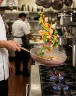 Falez 9-piece pink ceramic granite cookware set featuring a fry pan tossing vegetables, a shallow casserole, and three pots with glass lids, arranged on a kitchen counter and dining table.