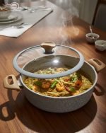 Falez 9-piece grey granite non-stick cookware set displaying a frying pan, shallow casserole, and deep pots with glass lids and wooden-style handles on a kitchen countertop.