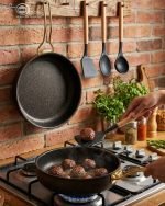 One More 12-piece black granite non-stick cookware set displayed on a wooden table, featuring gold handles, glass lids, a fry pan, shallow casserole, deep pots, and cooking spoons.