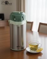 3-Liter Stainless Steel Airpot Thermos Flask with pump action dispenser and glass liner, displayed on a kitchen counter in Blue, Brown, and Green color options. Features callouts for rotating base, air-tight cap, and sleek handle.