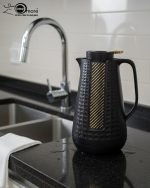 1 Litre vacuum flask thermos jug featuring a textured geometric design and gold accent strip. Shown in Off-White, Grey, and Black colors on a marble kitchen counter with callouts highlighting the glass interior, sleek handle, and air-tight cap.