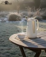 1 Litre vacuum flask thermos jug featuring a textured geometric design and gold accent strip. Shown in Off-White, Grey, and Black colors on a marble kitchen counter with callouts highlighting the glass interior, sleek handle, and air-tight cap.