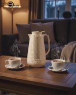 1 Litre vacuum flask thermos jug featuring a textured geometric design and gold accent strip. Shown in Off-White, Grey, and Black colors on a marble kitchen counter with callouts highlighting the glass interior, sleek handle, and air-tight cap.