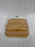 A chocolate cake with caramel drizzle sitting on a square bamboo wooden base, covered by a clear acrylic dome with a crystal handle on a white marble kitchen countertop.