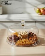 A versatile 2-in-1 clear acrylic serving set shown in two views: one as a divided platter filled with dates, raisins, and walnuts under a dome lid, and the other as a pedestal stand holding a layered chocolate cake on a marble kitchen counter.