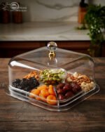 A versatile clear acrylic 2-in-1 serving dish shown in two uses: one as a divided tray filled with assorted dry fruits, nuts, and apricots, and the other as a pedestal cake stand holding a caramel-drizzled cake under a protective dome.