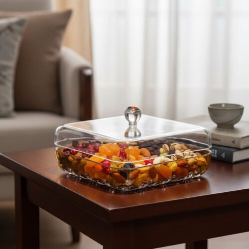 A square acrylic dry fruit dish with a gold-plated rim and crystal-patterned base, featuring a clear lid with a round knob, filled with assorted nuts and apricots on a wooden table.