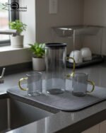 A person carrying a black serving tray with a modern smoke-grey glass water pitcher and six matching mugs with amber handles, filled with ice water in a bright, minimalist living room.