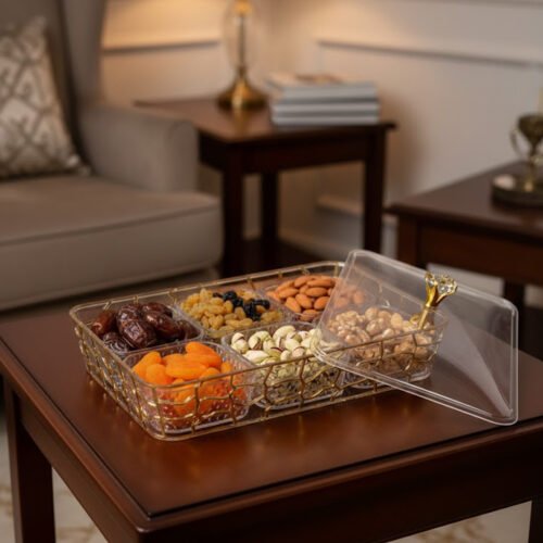 A 6-compartment clear acrylic dry fruit tray containing dates, apricots, almonds, pistachios, and raisins, held in a gold mesh frame on a dark wooden side table.