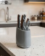 A modern 6-piece kitchen knife set by OneMore featuring grey ergonomic handles with copper accents, displayed both inside a sleek grey storage block on a granite countertop and laid out on a white surface to show the stainless steel blades and matching kitchen shears.