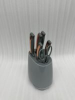 A modern 6-piece kitchen knife set by OneMore featuring grey ergonomic handles with copper accents, displayed both inside a sleek grey storage block on a granite countertop and laid out on a white surface to show the stainless steel blades and matching kitchen shears.