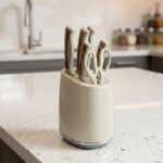 A 6-piece kitchen knife set featuring cream-colored ergonomic handles with rose gold accents, displayed both inside a matching compact storage block on a granite countertop and laid out on a white surface to show the different stainless steel blades and kitchen shears.