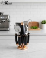 A 7-piece luxury kitchen tool set featuring black and rose gold handles, including a whisk, pizza cutter, and grater, arranged on a matching black and gold rotating stand on a marble countertop.