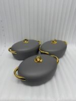 A set of three modern, oval-shaped insulated hotpots with gold handles and knobs, shown in chocolate brown, grey, and white colors on wooden and marble dining surfaces.