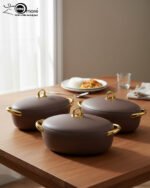 A set of three modern, oval-shaped insulated hotpots with gold handles and knobs, shown in chocolate brown, grey, and white colors on wooden and marble dining surfaces.