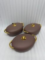 A set of three modern, oval-shaped insulated hotpots with gold handles and knobs, shown in chocolate brown, grey, and white colors on wooden and marble dining surfaces.