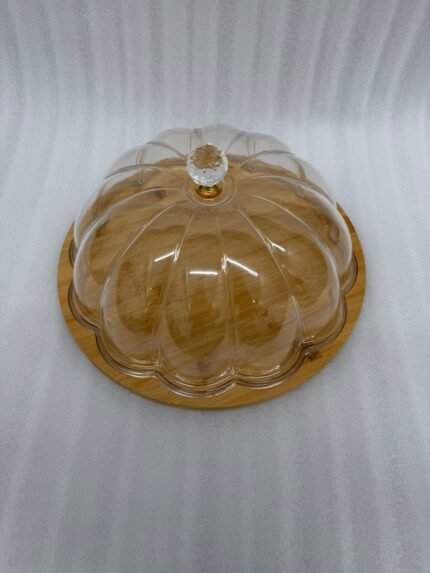 Two round cake stands of different sizes on a white kitchen counter, featuring natural wooden bamboo bases and clear ribbed acrylic dome covers with decorative crystal knobs, containing fruit cakes.