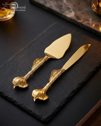 A luxury gold-plated cake knife and server set with textured ginkgo leaf handles resting on a black slate serving board.