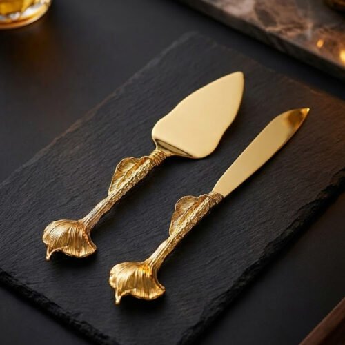 A luxury gold-plated cake knife and server set with textured ginkgo leaf handles resting on a black slate serving board.