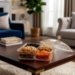 A square 4-compartment clear acrylic dry fruit box featuring a golden geometric lattice exterior and a transparent lid with a crystal knob, filled with assorted nuts and dried fruits on a wooden table.