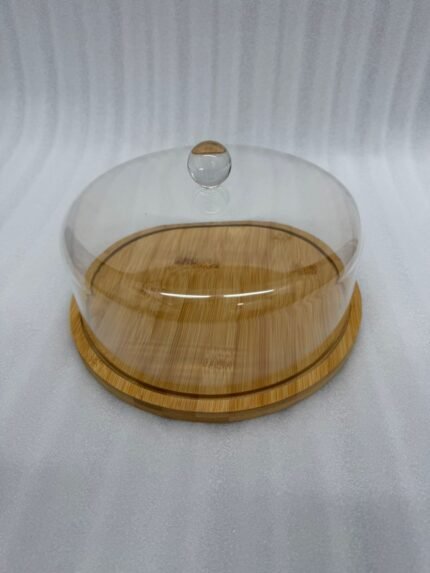 A white frosted layer cake displayed on a natural wooden bamboo base, covered by a clear circular acrylic dome with a round glass-like knob on a white marble kitchen counter.